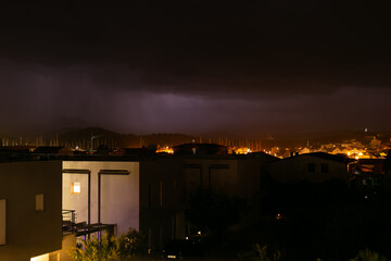 A coastal town illuminated by warm lights, with dark storm clouds gathering overhead. The distant horizon glows faintly as a storm approaches, creating a dramatic contrast between light and dark.