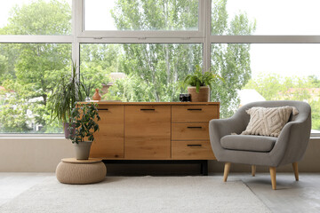 Wooden chest of drawers with grey armchair and houseplants near window in living room