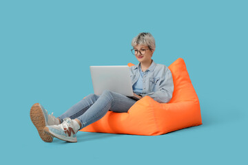 Portrait of beautiful young woman with laptop sitting on bean bag chair against blue background