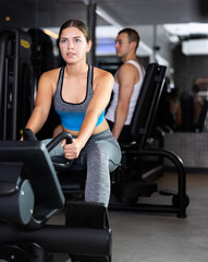 Fototapeta premium During workout, girl sits on simulator and pulls holder with her hands with effort. She does athletic exercises to give his muscles strength and endurance