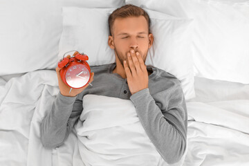Young man yawing and holding clock in bed at home. Insomnia concept