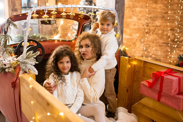 Mother with her little children near Christmas trees and lights in red car pickup trailer