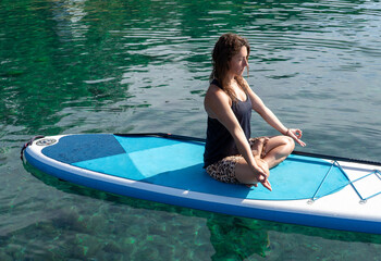 Young woman balancing on a stand-up paddleboard while practicing yoga on a serene lake, surrounded...