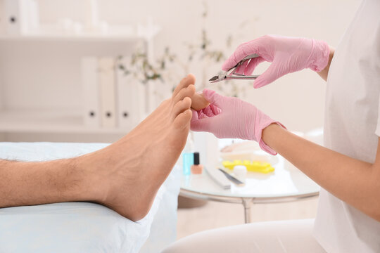 Beautician cutting cuticle off feet of young man on couch in spa salon, closeup