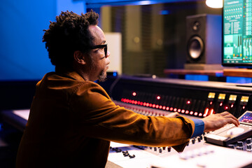 African american record producer adjusting sound settings on control board desk, editing tunes in professional studio. Music engineer working on mixing console to edit music, pushing faders.