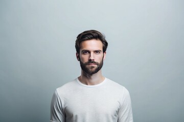 Obraz premium A young man with a thoughtful expression stands confidently against a light gray background, his casual white shirt accentuating his striking features and deep gaze. Generative AI