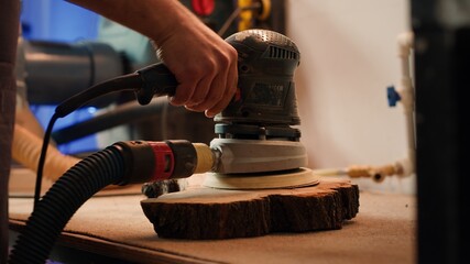 Cabinetmaker at workbench using orbital sander with fine sandpaper on lumber to achieve smooth finish. Woodworking specialist uses angle grinder on wood for professional results, close up, camera A