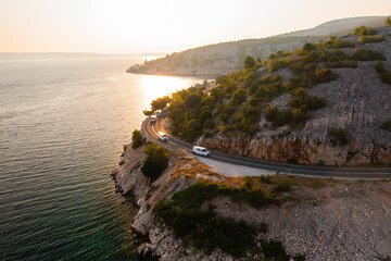 Scenic coastal drive along rocky shoreline at sunset with vehicles on winding road. 