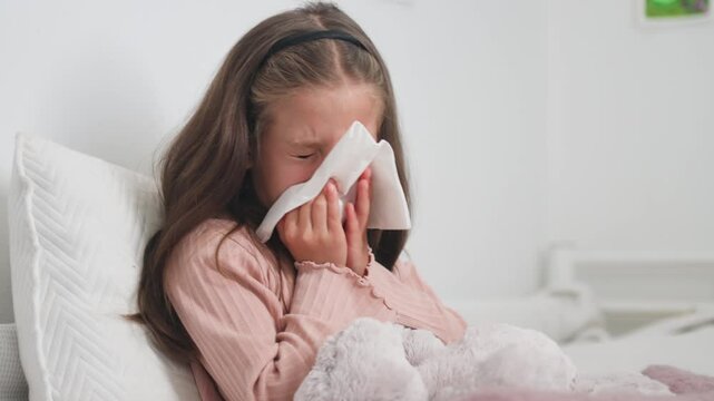 Sick girl sneezing snot into tissue paper in bedroom at home. Female child blowing runny nose is suffering from cold and flu. 