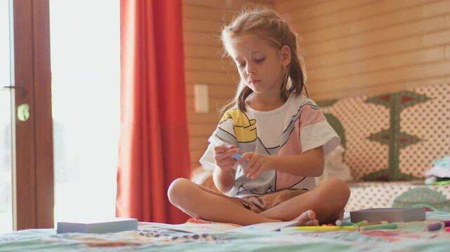 Cute girl painting on paper with felt tip pen while sitting on bed. Artistic female child is making art at home. 
