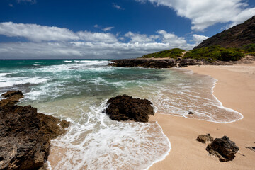 A sunny day with few clouds at the remote and pristine Hawaiian Monk Seal Beach in Oahu’s Ka’ena Point Beach Park, with waves stretching up the beach and lava rocks in the foreground