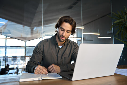 Busy professional young business man manager worker wearing earbuds working in office elearning or having hybrid meeting learning using laptop computer watching webinar writing notes sitting at desk.
