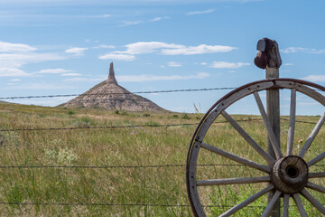 A rustic wagon wheel in the foreground with Chimney Rock rising majestically against a clear blue sky in Nebraska’s prairie landscape