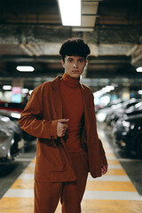 Young man in a brown suit stands confidently in a parking garage, surrounded by cars and bright overhead lights