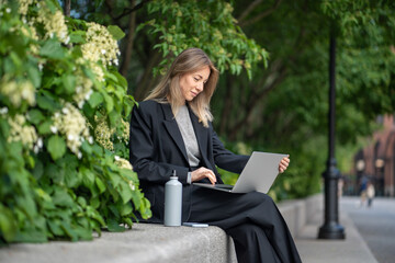 Pleased woman freelancer sits outdoor with portable computer busy job, performs project duties....