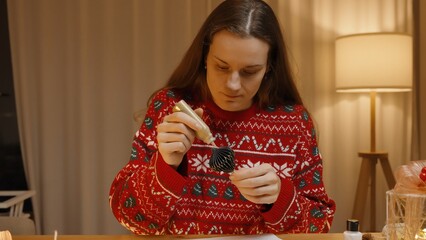 Applying Gold Glitter Glue to a Christmas Ornament, Focused and Wearing a Festive Sweater with a Holiday Background.