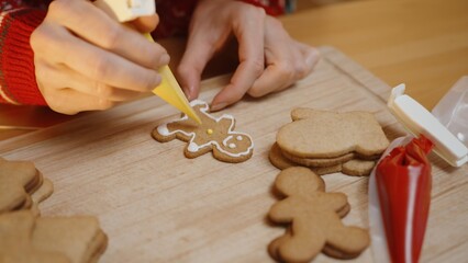 Hands Continue Decorating a Gingerbread Man Cookie With Yellow Icing on a Wooden Board, Surrounded by Other Plain Cookies and Icing Bags.