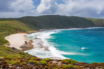 Sharp Point Lookout in Western Australia, featuring a rocky coastline, turquoise ocean waves, and a sandy beach. Dense green vegetation covers the hillsides under a cloudy sky.