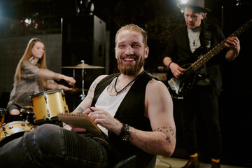 Medium portrait of happy young Caucasian male rock musician sitting on chair with notepad in hands smiling at camera