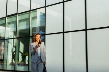 Business woman strolls confidently outdoors in smart attire, checking her messages against the backdrop of a modern glass building on a sunny day