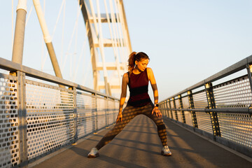 Dynamic morning workout on a sunlit bridge showcasing a young woman stretching and embracing fitness in an urban environment