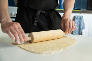 Hands of a man baking dough with rolling pin on white table