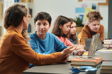 Fototapeta premium Portrait of Middle Eastern young boy talking to teacher during computer class in school with group of children copy space