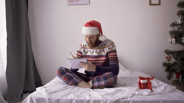 Young man wearing christmas sweater and santa hat in bedroom sitting on bed writing notes in notebook