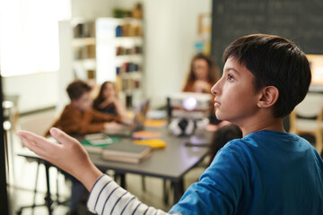 Side view portrait of young boy pointing at blackboard while answering questions or presenting project in school classroom copy space
