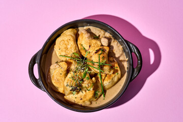 A close-up of a cast iron skillet filled with golden-brown, baked varenyky. The varenyky are garnished with fresh herbs on pink background.