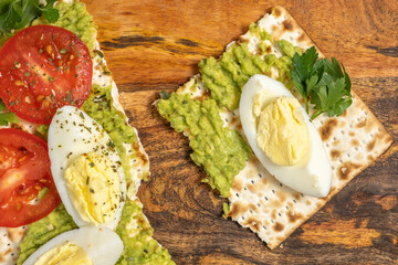 Matzah toast with creamy avocado, fresh tomatoes, boiled eggs on wooden background for Jewish holiday Pesach.