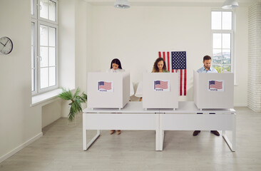 Group of young confident American citizens voters standing in a row at vote center with USA flags in voting booth. People voting at polling station on election day. Democracy concept.