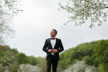 A man in a black suit and white shirt stands in a field of trees. He is wearing a bow tie and he is looking up at the sky. The scene is peaceful and serene
