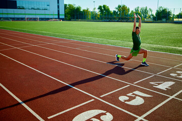Young fit man, athlete stretches at starting line, preparing for race on sunlit track, with lush green field in background. Concept of sport, tournament, motivation, workout. Ad