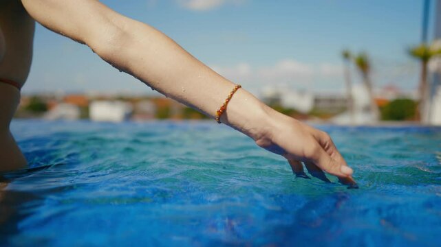 Woman touching water of swimming pool, closeup view of female hand, relaxation. Traveling and tourism in summertime, bright sun and blue water, enjoying warm water for refreshing in thermal baths