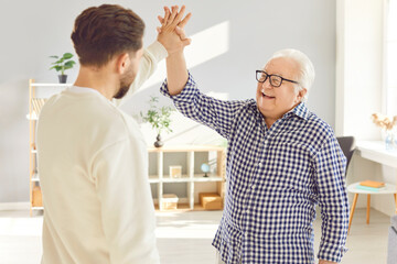 Fototapeta premium Happy smiling senior old parent giving high five to adult son when they meet in living room. Young man visiting elderly father greeting him at home. Mature parents, family and fathers day concept.
