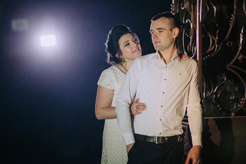 A man and woman are standing together in the dark. The man is wearing a white shirt and the woman is wearing a white dress. They are both smiling and looking at the camera