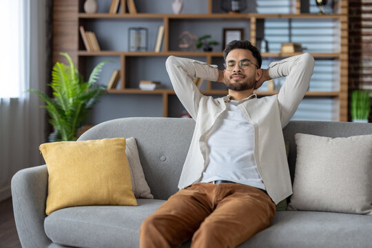 Young man relaxing on comfortable sofa at home, enjoying peaceful moment with hands behind head