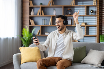 Excited young man listening to music with headphones and phone in comfortable living room atmosphere