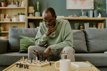 Young African American man thinking over tactics while solving chess problem in living room at home