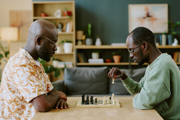 Side view shot of two African American brothers sitting at wooden table in front of each other and playing chess