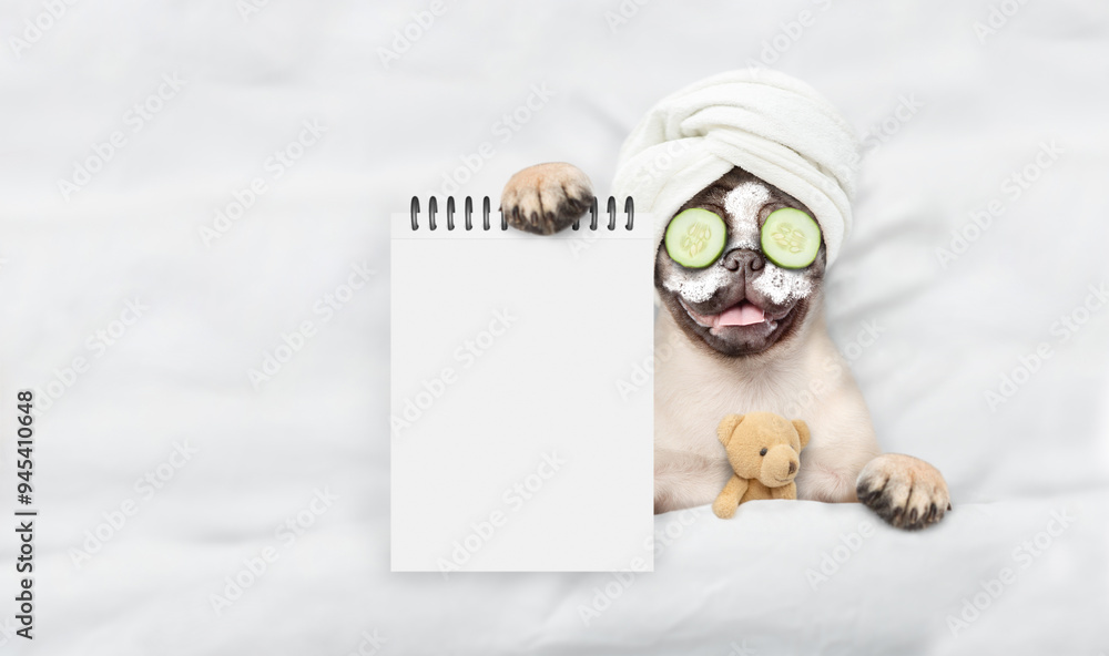 Aufkleber Funny Pug puppy with towel on it head and with pieces of cucumber on it eyes relaxing on the bed at home and showing empty list. Top down view. Empty space for text