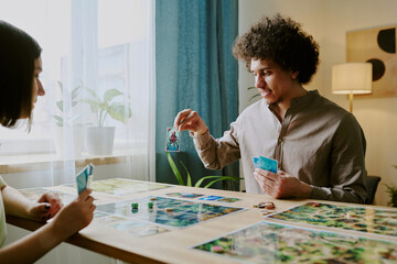 Young biracial man showing his card to girlfriend while playing modern fantasy board game together at home
