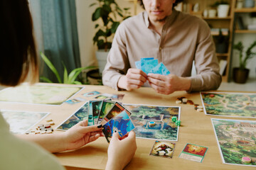 Unrecognizable young woman and her friend sitting at table in front of each other with cards in...