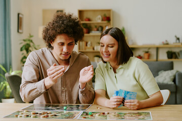 Medium shot of young ethnically diverse couple throwing dice while playing modern tabletop game in...