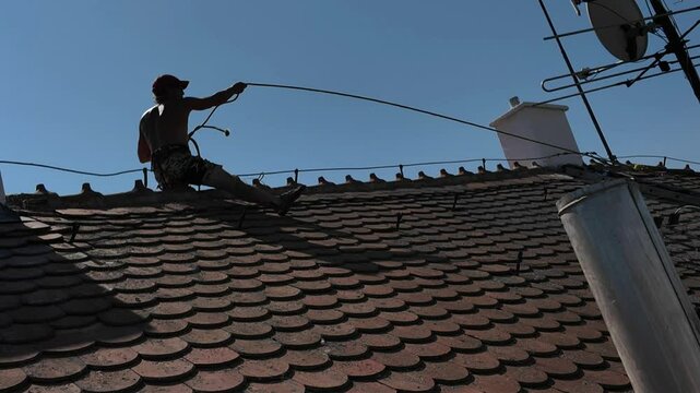 Worker on the ridge of an old clay tile roof. A laborer repairs the roof. Workman on roof with safety rope and special harness. Concept of construction work and working at heights