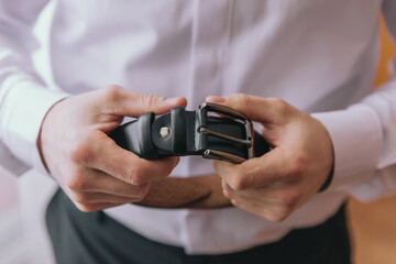 A man is holding a belt in his hands. The belt is black and has a silver buckle. The man is wearing a white shirt and black pants