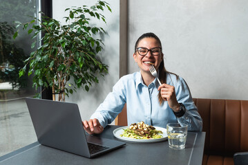 Young business woman working remotely from cafeteria during lunch break. Female office worker using laptop computer while she eating at restaurant to work or watching funny content on internet.