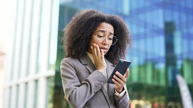 African American businesswoman in gray suit looking shocked while reading bad news on phone in front of modern office building. Black woman showing concern and disbelief. Worry and stress. Close up