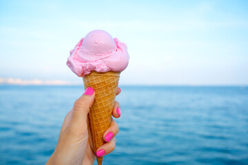 Pink ice cream in the girl’s hand with pink manicure  against sea backdrop 